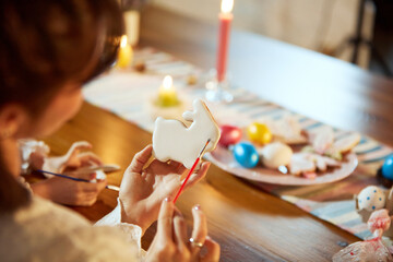 Focus on female hands, mother sitting at table at home and decorating baked gingerbread for Easter holiday celebration. Concept of holiday, Easter, family, motherhood and childhood