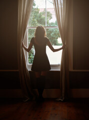 Woman, curtain and morning by window for sunlight, vitamin D or sunshine in bedroom at home. Rear view of female person standing by blinds or glass in casual clothing and looking out in neighborhood