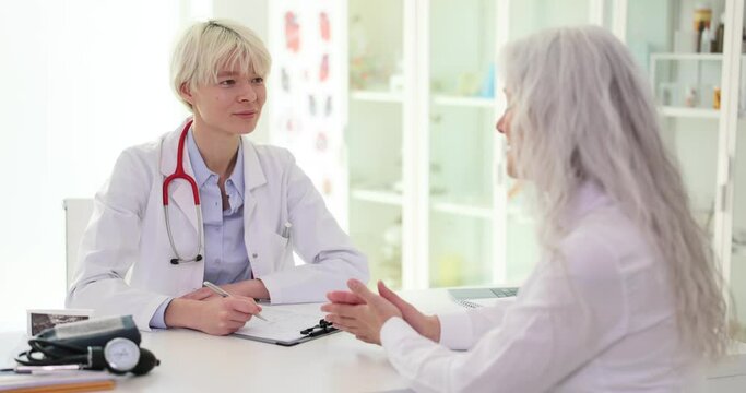 Female doctor listens, elderly woman talks about chronic diseases health complaints symptoms of ailment. Therapist takes notes fills out history form during patient visit