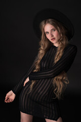 Portrait of blonde model exuding sensuality in relaxed pose on black background, looking at camera. Fashionista wears chic, wide-brimmed hat, black tight knit dress that shows off slender, petite body