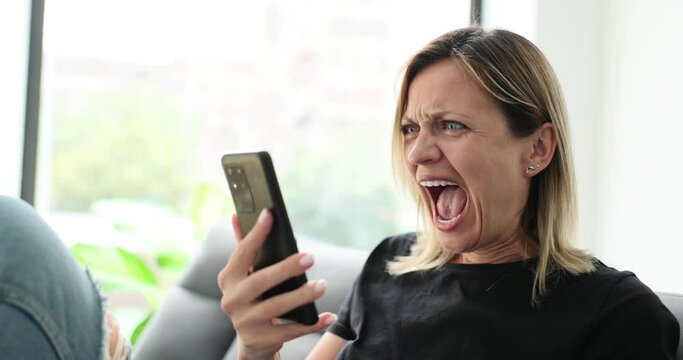 Angry woman yells at phone and looks at screen sitting on couch at home. Annoyed woman reading bad news and annoyed by spam