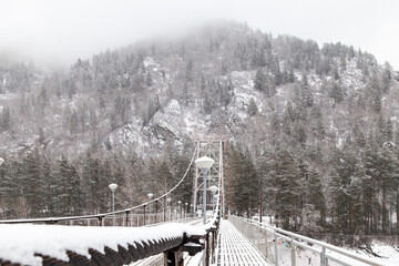 metal suspension bridge for cars and pedestrians in the Altai mountains in winter with snow and fog on the trees without people. Picturesque place winter landscape