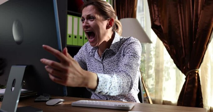 Irritated angry woman sitting at table next to computer and swears at broken monitor. Malware and malfunction and computer repair concept