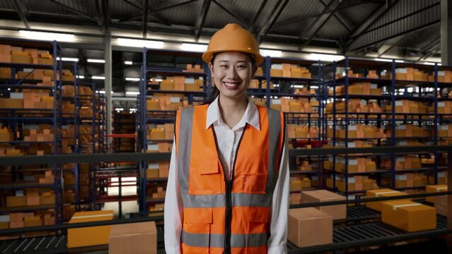 Asian Female Engineer With Safety Helmet Smiling To Camera While Standing In The Warehouse With Shelves Full Of Delivery Goods
