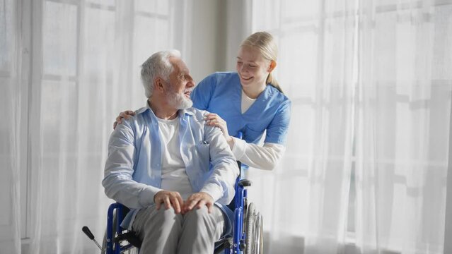 Social worker helps man in wheelchair. Woman nurse social welfare physical therapist looks after senior male, talking, providing medical care at home. Living with legs paralysis, special needs people.