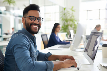 Confident Human Resources Agent Smiling Happily While Collaborating Online with Colleague. Portrait of younger man working at his computer in a modern office environment with shelves and plants in the