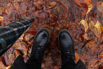 Top view of woman's feet in black winter boots standing in the rain on wet street in pile of fallen leaves, holding an umbrella