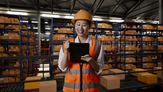 Asian Female Engineer With Safety Helmet Standing In The Warehouse With Shelves Full Of Delivery Goods. Taking Note On The Tablet And Looking Around In The Storage
