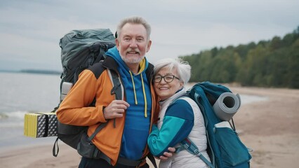Portrait senior couple travelers hikers carrying backpacks with touristic equipment standing on riverbank looking at camera. Man woman pensioners hiking together. Tourism, adventure, hiking concept.