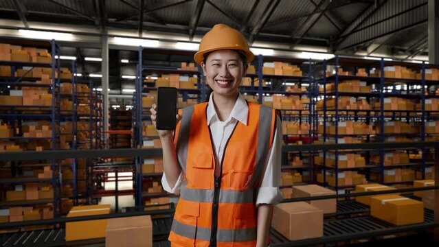 Asian Female Engineer With Safety Helmet Standing In The Warehouse With Shelves Full Of Delivery Goods. Smiling And Showing Black Screen Smartphone To The Camera In The Storage

