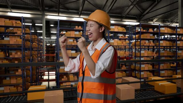 Side View Of Asian Female Engineer With Safety Helmet Standing And Screaming Goal Celebrating Working In The Warehouse With Shelves Full Of Delivery Goods
