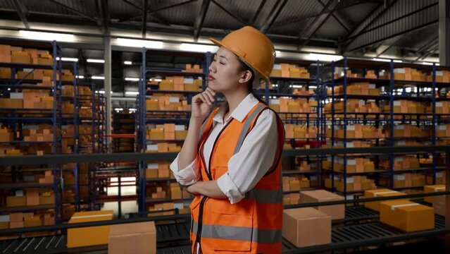 Side View Of Asian Female Engineer With Safety Helmet Standing In The Warehouse With Shelves Full Of Delivery Goods. Thinking About Something And Looking Around In The Storage
