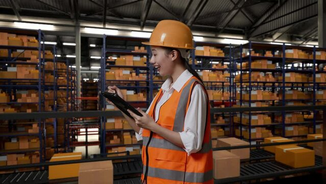 Side View Of Asian Female Engineer With Safety Helmet Standing In The Warehouse With Shelves Full Of Delivery Goods. Taking Note On The Tablet And Looking Around In The Storage

