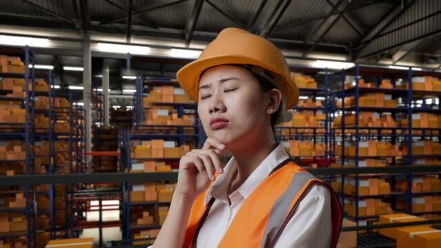 Close Up Side View Of Asian Female Engineer With Safety Helmet Standing In The Warehouse With Shelves Full Of Delivery Goods. Thinking About Something And Looking Around Then Raising Her Index Finger 