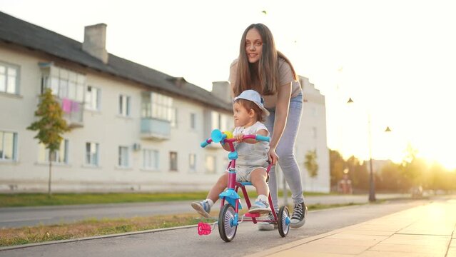 mother teaches daughter to ride. happy family kids dream concept. young mother teaches her daughter to small tricycle along the city center road. young mother teaches her little daughter bicycle