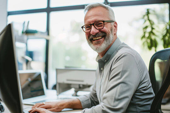Portrait of a cheerful, bearded, and bespectacled older man with white hair, engaging with the camera with a bright smile while working at his computer in a sunny, modern office environment.