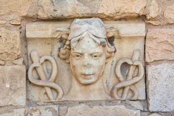 Stone-carved head of Hermes with wings rising from his hair and caduceus on both sides on the ruins...