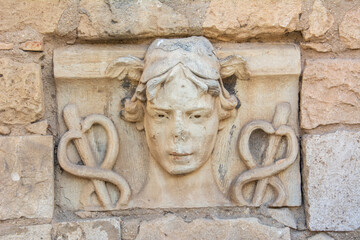 Stone-carved head of Hermes with wings rising from his hair and caduceus on both sides on the ruins of an ancient Roman time wall in Spain