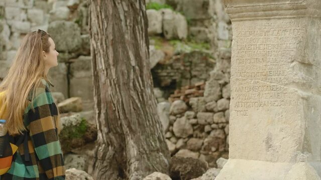 The female traveler examines an ancient Greek stone column with inscriptions carved into it, against the backdrop of ruins in the ancient city.
