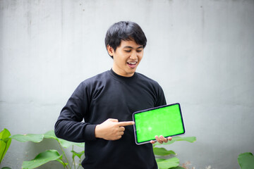 Young Asian man with a black sweater holding and pointing at an iPad tablet green screen mockup