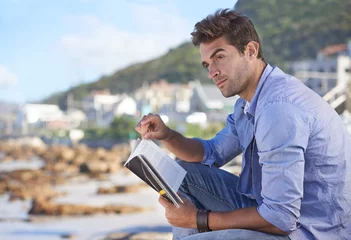 Poster Beach, book and young man reading for knowledge or relaxing hobby on vacation or holiday. Travel, outdoor and handsome confident male person enjoying story or novel on weekend trip or adventure. © Hover/peopleimages.com
