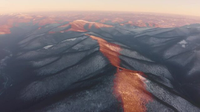 drone flies over the peaks of the mountainous Carpathian region with a winter color pattern of snowy beech trees on the tops, river valleys painted with sunset flames