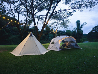 Outdoor camping tent with tarp on grass courtyard and warm night light under dark blue sky twilight time