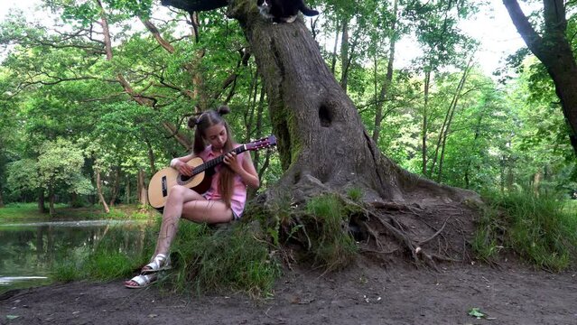a girl in pink clothes with long hair sits on a tree and plays the guitar, the cat is next to the girl and runs away to the side