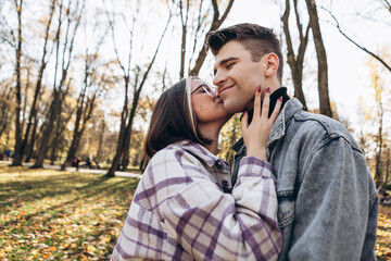 Heterosexual caucasian young loving couple walking outside in the city park in sunny weather, hugging smiling kissing laughing spending time together. Autumn, fall season, orange yellow red leaves
