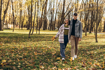 Fototapeta premium Heterosexual caucasian young loving couple walking outside in the city park in sunny weather, hugging smiling kissing laughing spending time together. Autumn, fall season, orange yellow red maple leav