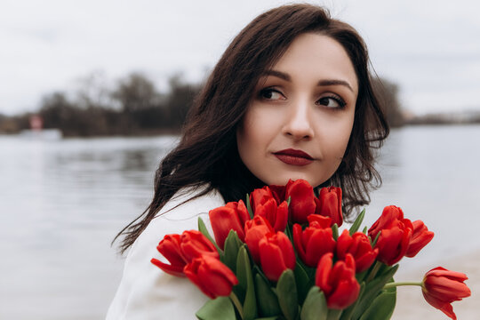 Attractive brunette woman walking on the beach shore in moody cloudy windy weather with bouquet of red tulips flowers, dressed in white suit jacket. International Women`s Day 8th March concept