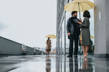 Obraz premium Business colleagues sharing an umbrella on a rainy city day with reflections.
