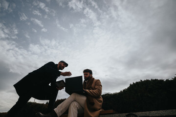 Contemplative men in coats discussing work on a laptop while sitting on a park bench under a...