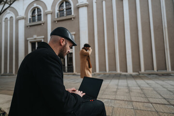 Fototapeta na wymiar A focused man in a coat and hat works on a laptop outdoors, with a blurred woman walking in the background by architectural columns.
