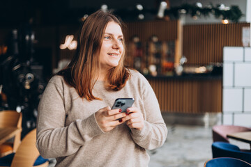 Young happy woman in glasses smiling and using smart phone in a cafe. Person using smartphone indoors. Online education, order, working or shopping concept