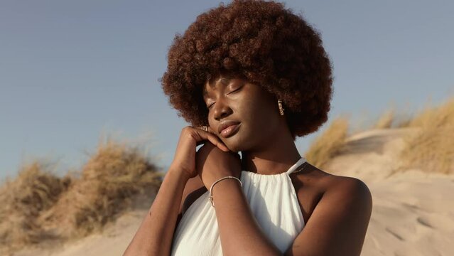 Elegant African woman with a natural afro hairstyle peacefully posing in sandy dunes, embracing the sunlight with eyes closed.