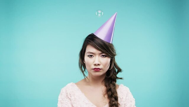 Woman, party and serious portrait in studio for celebration, achievement or new year on a blue background. Young asian person with bored face in birthday hat and bubbles in air for event or news