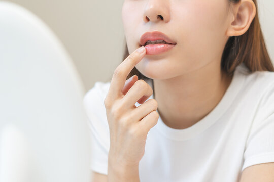 Lip care concept, worried asian young woman finger touching lips in front of mirror after applying lipstick balm on dry mouth from natural beauty product, skincare routine, makeup and cosmetics.