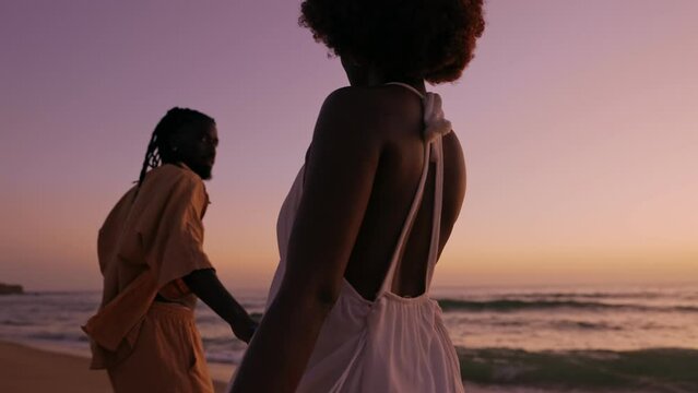 Silhouetted image of a young African couple in stylish attire walking along the beach during a beautiful sunset, displaying affection and togetherness.