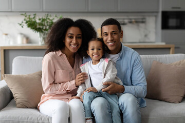 Cheerful black family of three people hugging sitting on couch at home, posing for photo and...