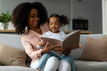 Caring young black woman reading book aloud to her cute little daughter, enjoying spending time together at home sitting on sofa