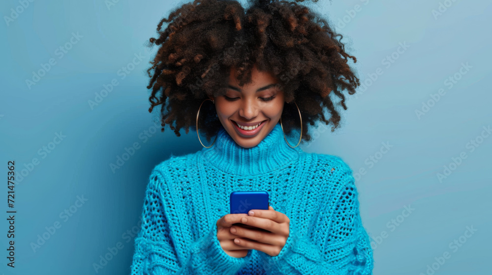 Poster young woman with a vibrant afro hairstyle is smiling and looking at her smartphone, wearing a cozy turquoise turtleneck sweater against a matching turquoise background