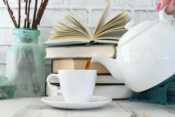 Pouring a cup of tea from a teapot with a stack of books and pastel spring accessories in the background. 