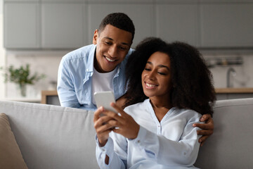 Positive African American couple sitting on couch, using smartphone, woman sharing social media...