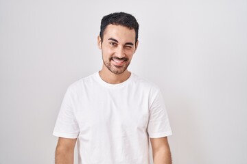 Obraz premium Handsome hispanic man standing over white background winking looking at the camera with sexy expression, cheerful and happy face.