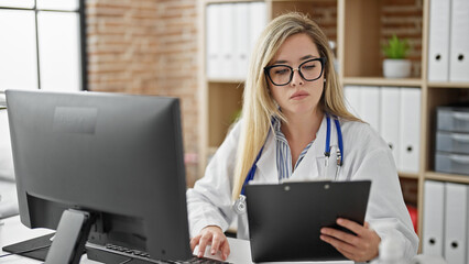 Young blonde woman doctor using computer reading medical report at clinic