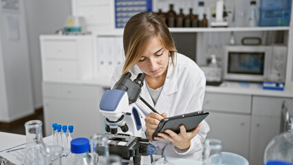Attractive young blonde scientist, close-up portrait of her concentrated work on analysis and research in the lab, using microscope and writing on the touchpad