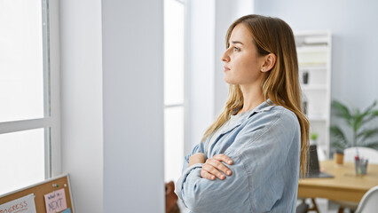 Young blonde business woman stands, arms crossed, looking through office window, ready to conquer...