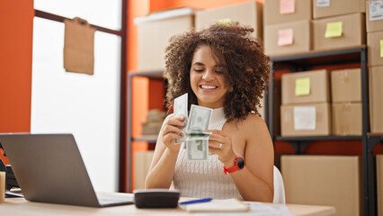 Young beautiful hispanic woman ecommerce business worker using laptop counting dollars at office