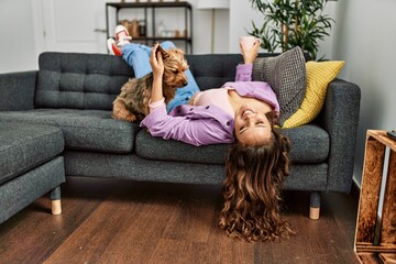 Young beautiful hispanic woman lying on sofa with dog holding cup of coffee at home - obrazy, fototapety, plakaty